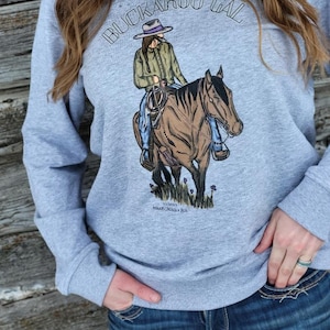 Könnte beinhalten: Ein graues Sweatshirt mit dem Text "BUCKAROO GAL" in schwarzen Buchstaben. Ein Cartoon-Bild einer Frau, die ein Pferd reitet, ist mittig auf dem Sweatshirt platziert. Die Frau trägt ein grünes Hemd, blaue Jeans und einen braunen Hut. Das Pferd ist braun mit einer schwarzen Mähne und einem schwarzen Schweif.