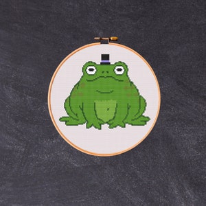 May include: A green frog wearing a black top hat with a purple band sits on a white background. The frog is embroidered on a round piece of fabric.