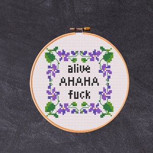 Puede incluir: Un aro de bordado blanco con un diseño de punto de cruz con flores moradas y el texto "alive AHA HA FUCK".