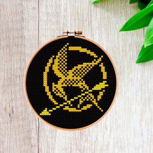 May include: A black and gold embroidery hoop featuring a cross-stitch design of the Mockingjay symbol from the Hunger Games. The symbol is a stylised bird with an arrow through it.
