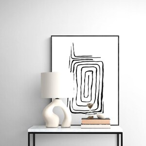 Puede incluir: Impresión de arte abstracto en blanco y negro con líneas en un marco negro sobre una pared blanca encima de una lámpara de mesa blanca y una pila de libros sobre una mesa blanca.
