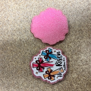 May include: Two decorative, glittery pink coasters. One is a solid pink shape. The other has three crosses with bows in red, blue, and orange, with the text "He is Risen Matthew 28:6".