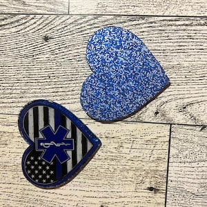 Op de afbeelding: Twee hartvormige decoraties. Eén is effen blauwe glitter. De andere heeft een blauwe lijnontwerp met een medisch symbool en elementen van de Amerikaanse vlag. Beide liggen op een licht houtnerfoppervlak.