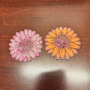 May include: Two sunflower-shaped resin decorations. One is pink with pink glitter, and the other is orange with glitter. The petals are outlined, and the centers are filled with glitter. These are likely decorative items.