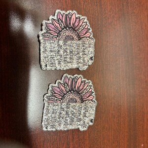May include: Two clear, glittery sunflower-shaped pins. Each pin features a pink and purple sunflower design above a rectangular section with the text: "She is clothed with strength & dignity and she laughs without fear of the future. Proverbs 31:25."