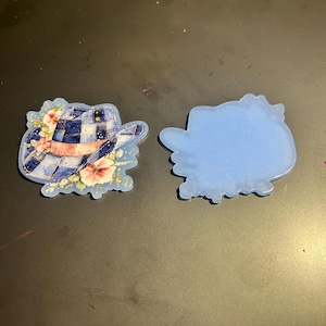 May include: Two resin cowboy hat-shaped coasters. One coaster features a blue and white plaid hat design with pink and white flowers. The other coaster is a solid light blue color. The coasters are on a dark surface.