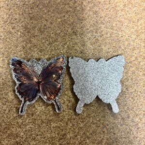 May include: Two butterfly-shaped embellishments. One features a detailed butterfly design with brown, black, and white wings, outlined in silver glitter. The other is a solid silver glitter butterfly shape.