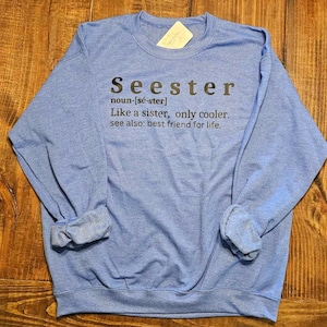 以下が含まれることがあります： 「Seester」の文字が黒でプリントされた水色のスウェットシャツ。「Seester」は「姉妹のように、ただもっとクール。また：生涯の親友」と定義されています。