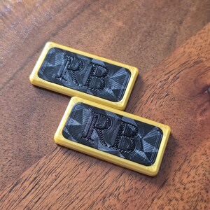 May include: Two rectangular, yellow-edged objects with a black and gray geometric pattern and the letters "RB" in black. The objects are resting on a wooden surface.