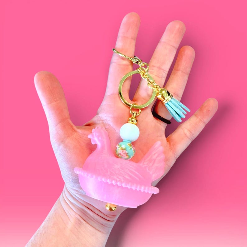 A hand holding a translucent pink 3D-printed hen-on-nest keychain with gold hardware, a floral bead, and a light blue tassel against a vibrant pink background.