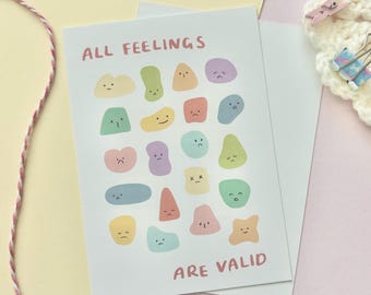 May include: A light blue card with the text "ALL FEELINGS ARE VALID" in pink lettering. The card is decorated with colorful cartoon shapes with different facial expressions.