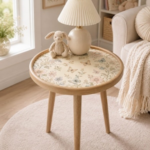 Puede incluir: Una pequeña mesa auxiliar redonda con estructura de madera clara y tres patas. La tapa presenta un diseño floral y de mariposas. Una lámpara beige con pantalla plisada y un conejito de peluche encima. La mesa tiene 61 cm de diámetro.