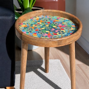 Może przedstawiać: Okrągły drewniany stolik pomocniczy z blatem w stylu mozaiki z kolorowymi geometrycznymi kształtami w odcieniach niebieskiego, pomarańczowego, różowego i zielonego. Stół ma trzy skośne nogi i podniesioną krawędź. Średnica stołu wynosi około 51 cm.