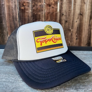 May include: White and black trucker hat with a Topo Chico logo on the front. The logo is yellow, red, and black with a white background. The hat has a black mesh back.