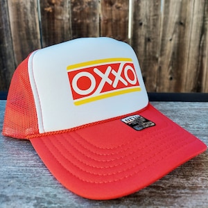Op de afbeelding: Een rode en witte truckercap met een geel en rood "OXXO" logo op de voorkant. De pet heeft een mesh achterkant en een rode klep.