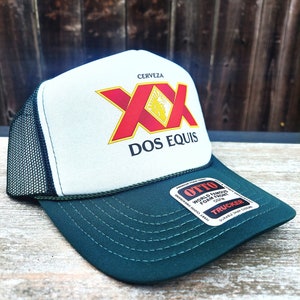 May include: A white and green trucker hat with a black mesh back. The front of the hat features a red and yellow logo that says "Cerveza XX Dos Equis". The back of the hat has a black label that says "Otto World Famous Foam Front OSFM Trucker Durable Snap Closure".