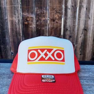 Op de afbeelding: Rode en witte truckercap met een geel en rood "OXXO" logo op de voorkant. De cap heeft een zwart "OTTO WORLD FAMOUS FOOD MART" label aan de zijkant.