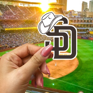 May include: A black and white sticker of the San Diego Padres logo with a white cowboy hat on top. The sticker is being held by a hand in front of a baseball stadium.