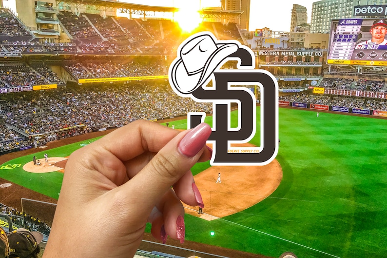May include: A black and white sticker of the San Diego Padres logo with a white cowboy hat on top. The sticker is being held by a hand in front of a baseball stadium.