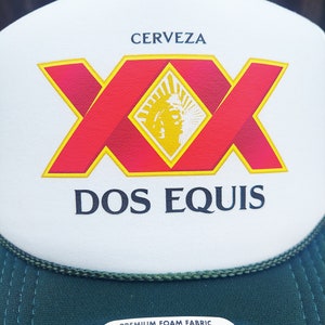 May include: A white and green trucker hat with a red and yellow Dos Equis Cerveza logo. The hat has a green mesh back and a black label that says "Premium Foam Fabric OTTO".