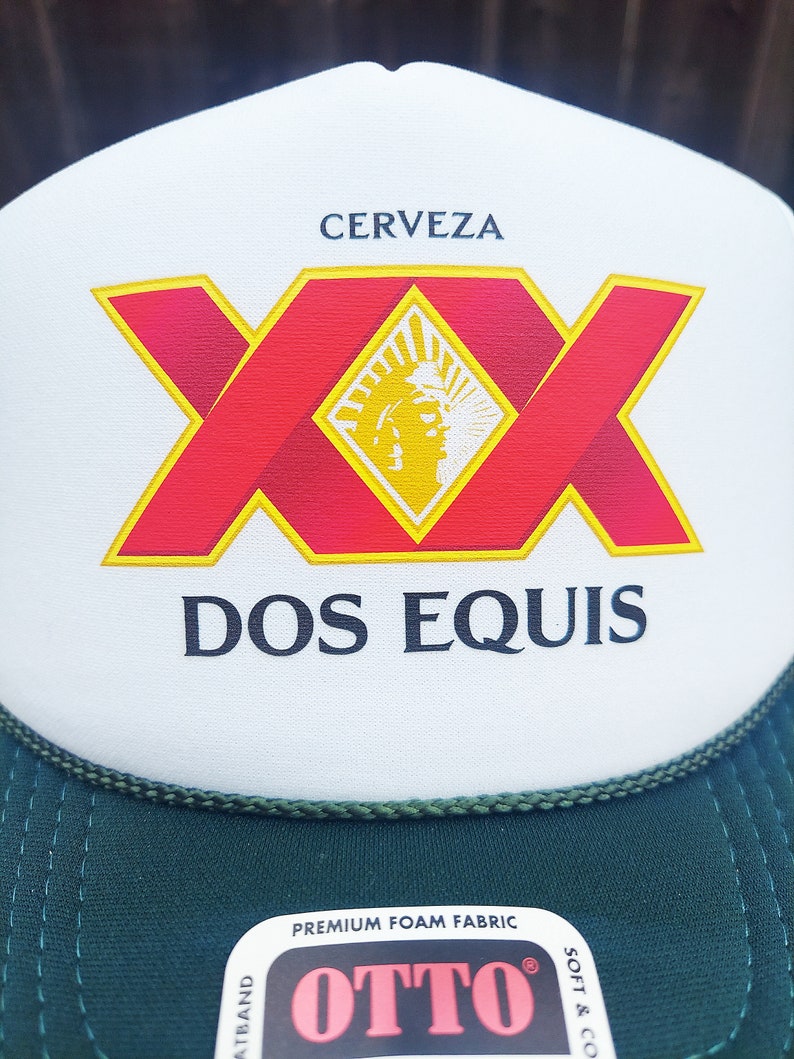 May include: A white and green trucker hat with a red and yellow Dos Equis Cerveza logo. The hat has a green mesh back and a black label that says "Premium Foam Fabric OTTO".