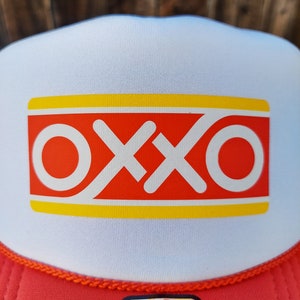 Op de afbeelding: Een witte en rode trucker-pet met een gele streep en het woord "OXXO" in rode letters. De pet heeft een rood mesh-achterpaneel en een zwart label met de tekst "OTTO WORLD FAMOUS FOAM FRONT OSFM".