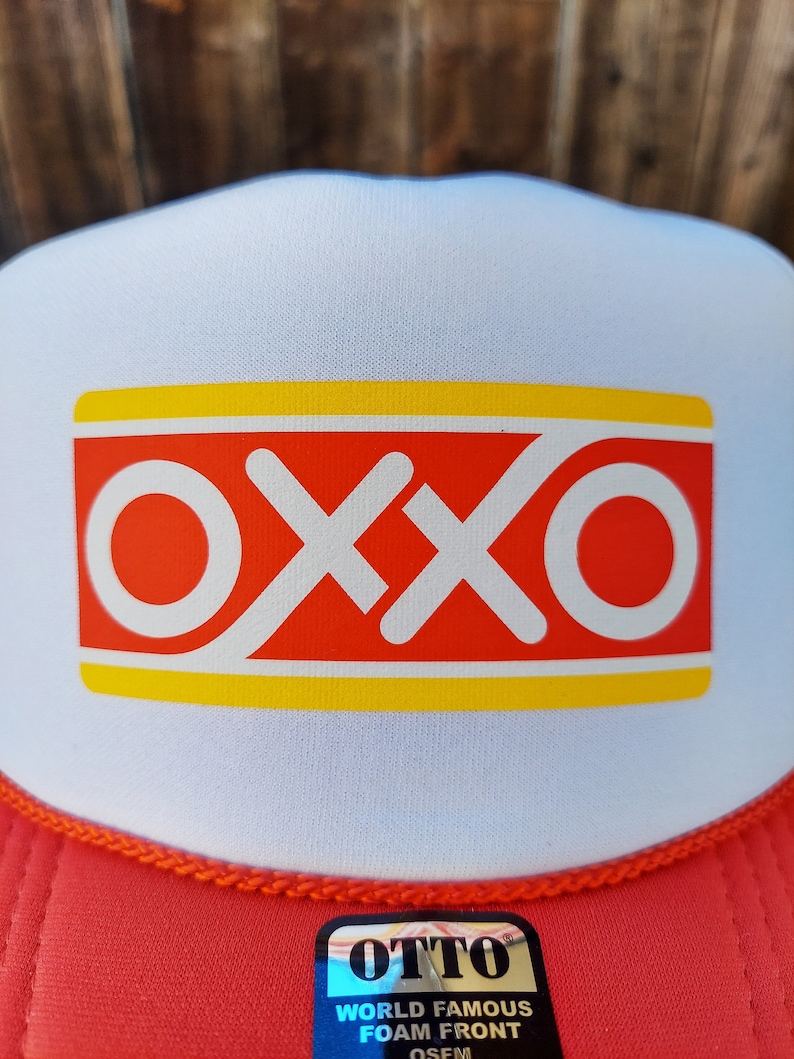Op de afbeelding: Een witte en rode trucker-pet met een gele streep en het woord "OXXO" in rode letters. De pet heeft een rood mesh-achterpaneel en een zwart label met de tekst "OTTO WORLD FAMOUS FOAM FRONT OSFM".