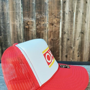 Op de afbeelding: Een rode en witte truckercap met een geel en rood "OK" logo op de voorkant. De pet heeft een rood mesh achterpaneel.