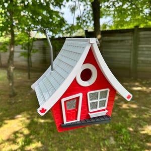 May include: A red and white birdhouse with a grey shingled roof, a round window, and a small chimney. The birdhouse has a door and a window with a small ledge. A maple leaf is on the side of the birdhouse.