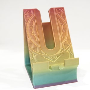 May include: A rainbow-colored phone stand with a decorative design. The stand is made of plastic and has a U-shaped cutout for holding a phone.