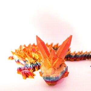 Puede incluir: Un dragón de origami colorido hecho de papel, con un esquema de colores rojo, naranja, amarillo, azul y verde. El dragón está enroscado en una posición de descanso.