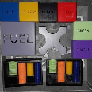 May include: A game board featuring a selection of coloured blocks, including blue, yellow, black, red, green, and purple. The blocks are arranged in a grid pattern and display the word "FUEL" on one of the blocks. The board also has a black and white X-shaped piece in the centre. There are also two black trays with rows of coloured tokens.