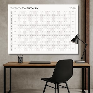 May include: A large, rectangular 2026 year calendar with the text "TWENTY TWENTY-SIX" at the top. The calendar is displayed on a wall above a wooden desk with a black chair and a black desk lamp.