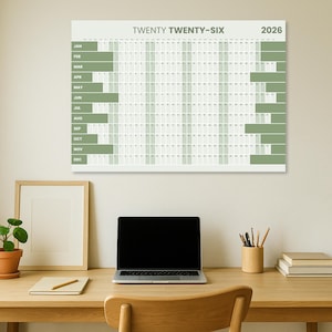 May include: A large wall calendar for the year 2026, with the text "TWENTY TWENTY-SIX" at the top. The calendar has a grid layout with months listed vertically and days horizontally. Green bars highlight specific periods. A laptop, desk accessories, and a framed picture are on a wooden desk.