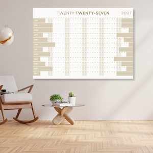 Könnte beinhalten: Ein großer, rechteckiger Wandkalender für das Jahr 2027. Der Kalender ist hauptsächlich beige und weiß, mit dem Text "TWENTY TWENTY-SEVEN" und dem Jahr 2027 oben. Die Monate sind vertikal links aufgeführt.