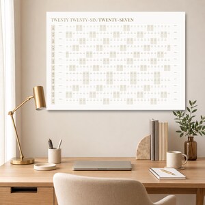 Könnte beinhalten: Ein großer, rechteckiger Wandkalender mit dem Text "TWENTY TWENTY-SIX/TWENTY-SEVEN" oben. Das Kalendarium ist beige, mit monatlichen Spalten und Daten. Der Kalender wird auf einem Schreibtisch mit einer goldenen Lampe, Büchern und einer Tasse angezeigt.