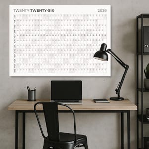 Könnte beinhalten: Ein großer weißer Kalender für 2026 mit dem Text "TWENTY TWENTY-SIX" ist an einer grauen Wand angebracht. Eine schwarze Schreibtischlampe, ein Laptop und Büromaterial stehen auf einem Holztisch. Vor dem Schreibtisch steht ein schwarzer Stuhl.