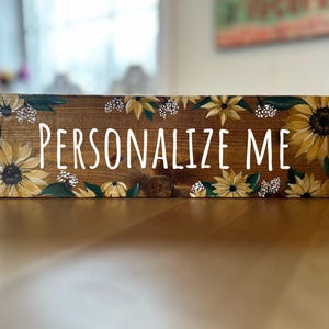 May include: A rectangular wooden sign with the words "PERSONALIZE ME" in white lettering. The sign is decorated with hand-painted sunflowers in shades of yellow and brown, with green leaves and white accents.