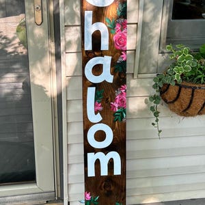 Könnte beinhalten: Vertikales Holzschild mit dem Wort "Shalom" in weißen Buchstaben. Verziert mit rosa Rosen und grünen Blättern. Der Hintergrund ist dunkles braunes Holz. Ein dekoratives Element.