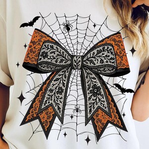 Puede incluir: Camiseta blanca con un diseño temático de Halloween. El gráfico presenta un gran lazo con encaje floral negro y blanco, bordes de encaje naranja y un fondo de telaraña. Murciélagos y arañas completan el diseño espeluznante.