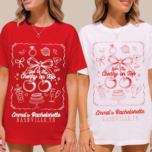 May include: Red and white t-shirts with a bachelorette party theme. Each shirt features a white graphic with cherries, a bow, and the text "She is The Cherry on Top" and "Emma's Bachelorette Nashville, TN."