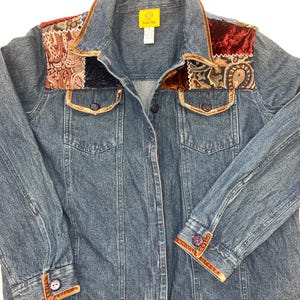 May include: A blue denim jacket with decorative patches of paisley and velvet. The jacket features brown trim along the collar, pockets, and cuffs. The cuffs and front closure have purple buttons. The jacket has a tag that says "Ruby Rd."