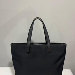 May include: A black tote bag with leather handles. The bag is rectangular with a structured design. The handles are attached to the top of the bag with leather accents. The bag is made of a durable fabric.