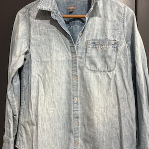 May include: Light blue denim shirt with a button-down front, a chest pocket, and long sleeves. The shirt is hanging on a wooden hanger. The shirt has a classic collar and buttoned cuffs.
