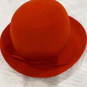 May include: A vibrant orange cloche hat with a rounded crown and a slightly upturned brim. The hat features a simple bow detail on the side. This stylish hat is made of a soft, felt-like material.