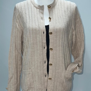 May include: A beige ribbed knit cardigan with a round neckline and gold-colored buttons. The long-sleeved sweater has two front pockets and button details on the cuffs. The cardigan is displayed on a mannequin.