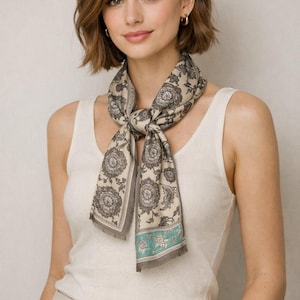 May include: A floral patterned scarf in cream, gray, and teal. The scarf is tied around the neck. The scarf has a floral design with a gray border and teal accents. The scarf is made of a soft, lightweight material.