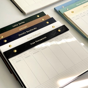 Puede incluir: Una pila de blocs de notas de escritorio personalizados con nombres en letras doradas. Los blocs de notas tienen un aspecto similar al cuero en varios colores, incluyendo verde, azul marino, negro y blanco. Cada bloc tiene un diseño de calendario semanal.