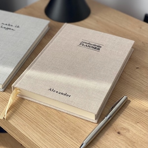 Peut inclure: Un planificateur de productivité recouvert de lin beige avec le nom "Alexander" imprimé sur la couverture. Un stylo argenté repose à côté du planificateur sur une surface en bois. Le planificateur a un signet en ruban de couleur crème.