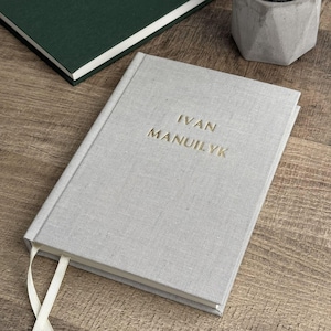May include: A light gray hardcover journal with the name "IVAN MANUILYK" embossed in gold lettering. A white ribbon bookmark extends from the journal. A dark green book and a small concrete pot are in the background.